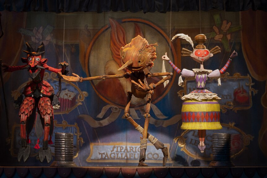 Three animated wooden-like puppets perform on a stage. In the centre is Pinocchio who is bowing with a smile.