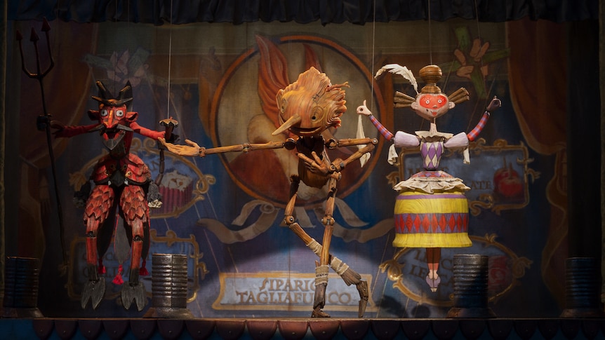 Three animated wooden-like puppets perform on a stage. In the centre is Pinocchio who is bowing with a smile.