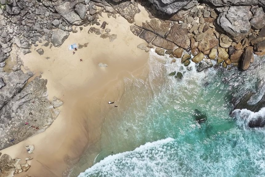 An aerial view of a small beach surrounded by rocks, with blue-green waves breaking on the shore.