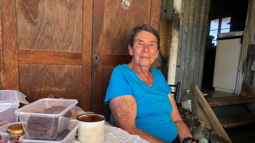 An older woman sits at her outside table with enamel cup and homemade fruit cake sharing recollections of earlier times