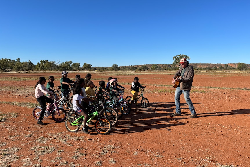 Man playing guitar to a group of children sitting on their bikes, all standing in the red dirt of outback Australia. 
