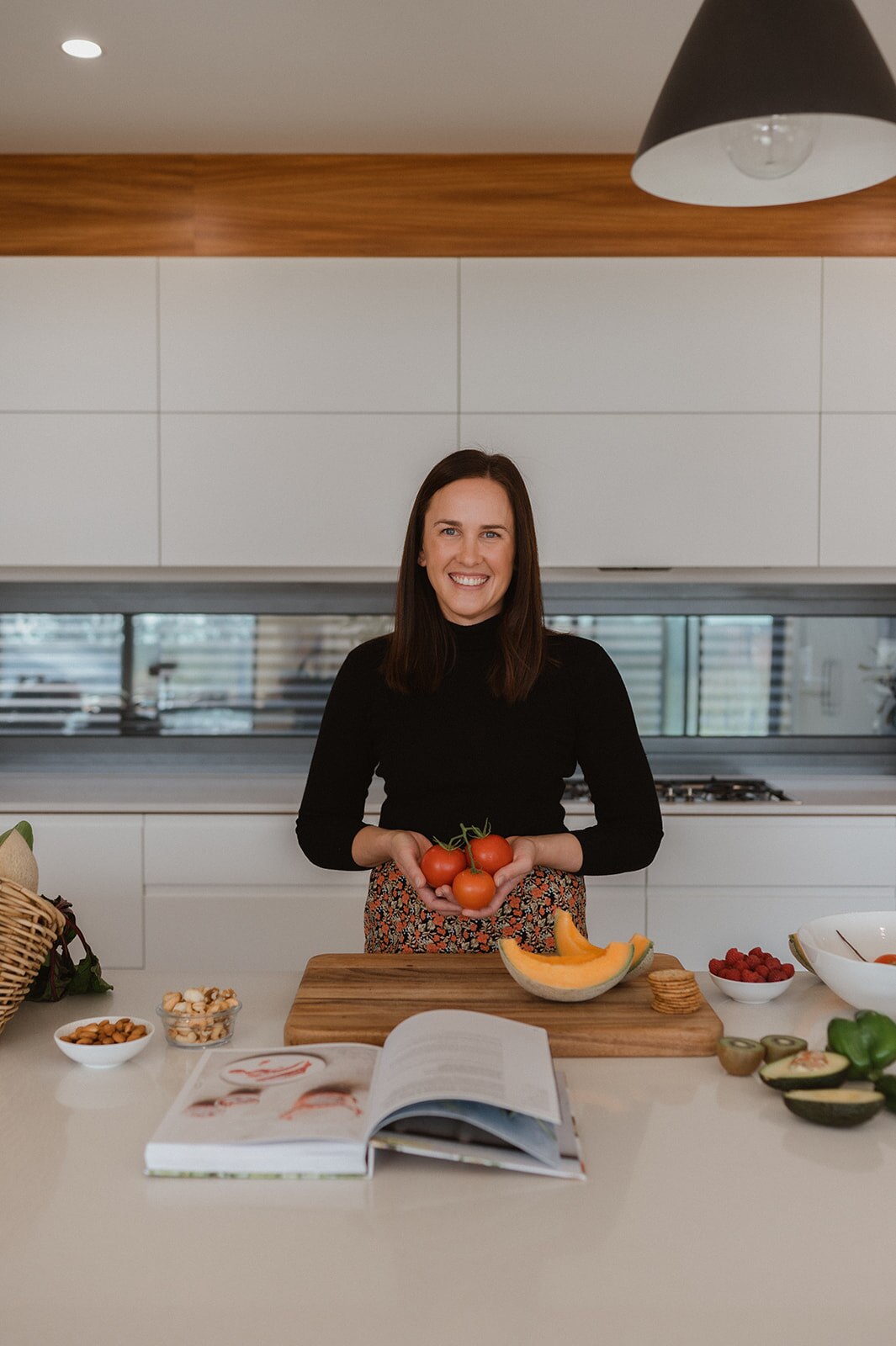 A woman smiles at the camera holding red tomatoes, she has long brown hair and a black top. she is in a white kitchen