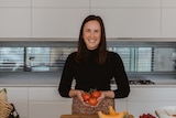 A woman smiles at the camera holding red tomatoes, she has long brown hair and a black top. she is in a white kitchen