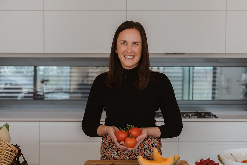 A woman smiles at the camera holding red tomatoes, she has long brown hair and a black top. she is in a white kitchen