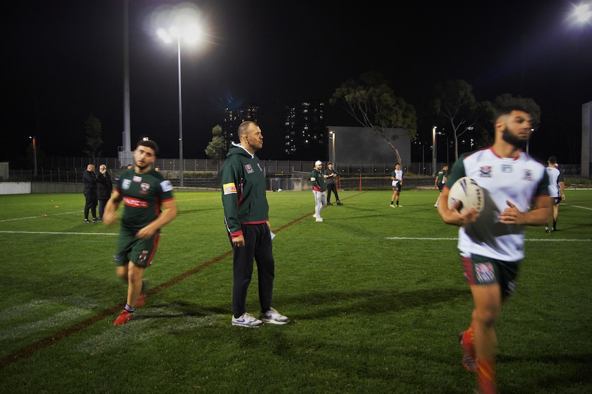 three men stand on a football field, one playing with a rugby ball.