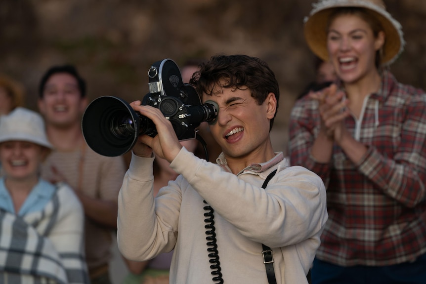 Teenaged white boy wears long sleeved white shirt and holds a black film video camera to his face while onlookers clap.