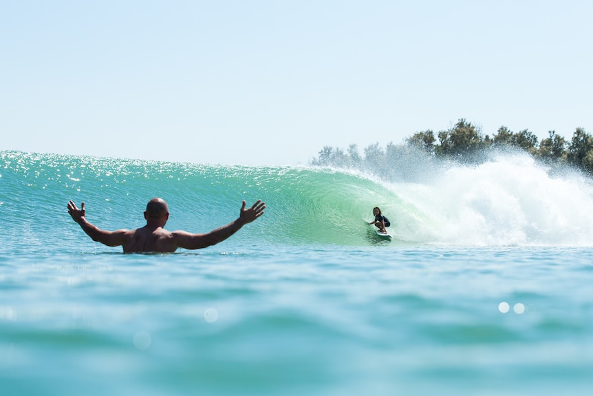 a surfer celebrating in the foreground as his son gets barreled in the backgrouns