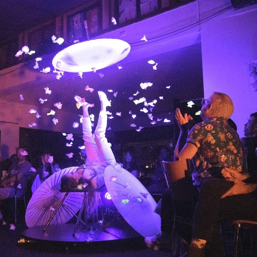A woman juggles a spinning umbrella on her feet, in a spotlight, while a live audience claps.