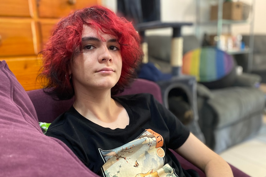 A pre-teen child with dyed red hair sits on a purple couch with a rainbow cushion in the background.