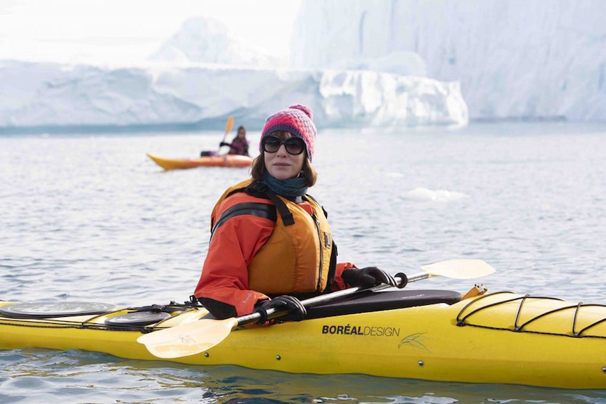 A woman wears sunglasses and beanie, holds kayak paddle and sits in yellow kayak in ocean in Antarctic-like landscape.