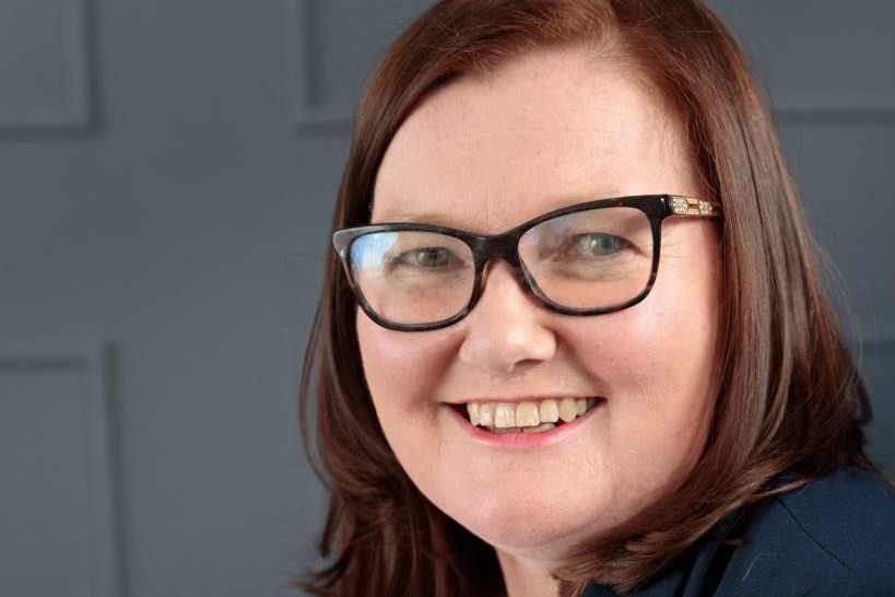 A headshot of a smiling Caucasian woman with red hair, black-rimmed glasses against a grey background.