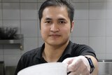 A close-up shot of a man holding out a plastic bowl in front of him while standing in a cafe kitchen.