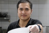 A close-up shot of a man holding out a plastic bowl in front of him while standing in a cafe kitchen.