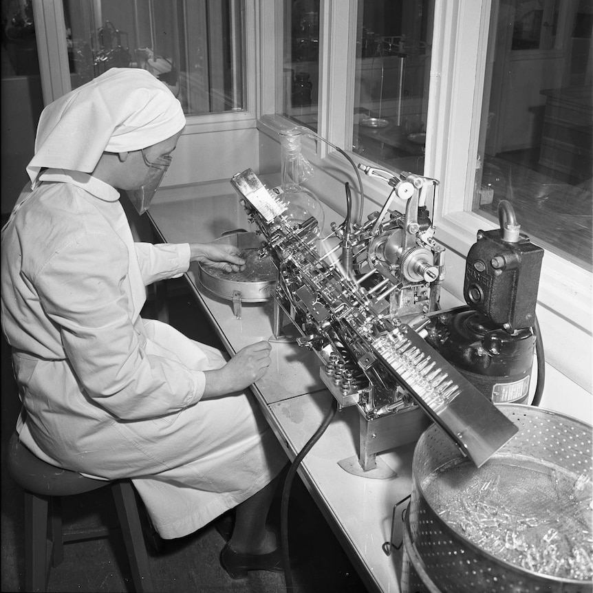 A woman in white medical uniform sits at a desk while packing pills via an elaborate metal machine.