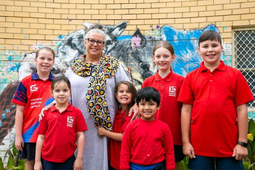 A woman with grey hair smiles with her arms around six children in school uniform.