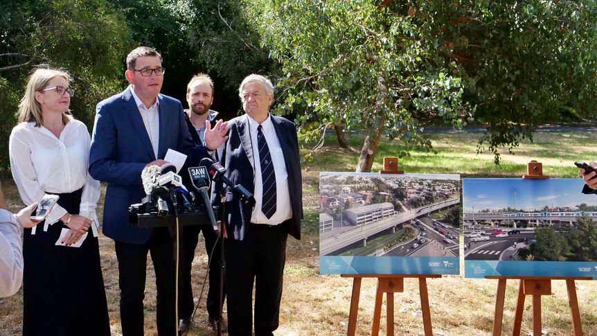 The Premier Daniel Andrews speaks into microphones near prints of artists impersonations of a elevated rail line.