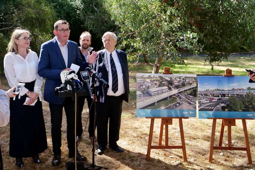 The Premier Daniel Andrews speaks into microphones near prints of artists impersonations of a elevated rail line.
