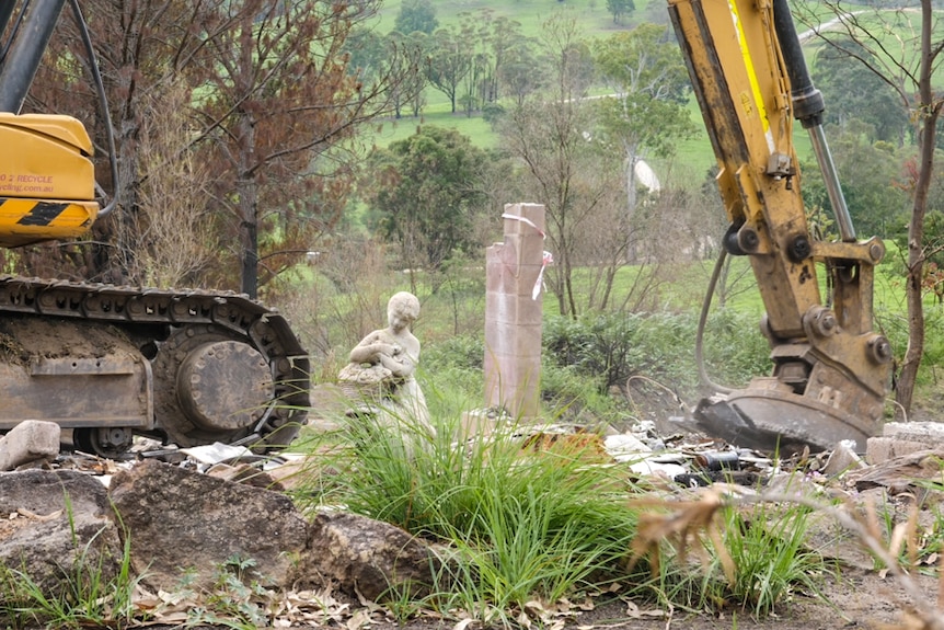 A statue of a young girl stands amongst the ruins of a burnt home as an excavator works around it.