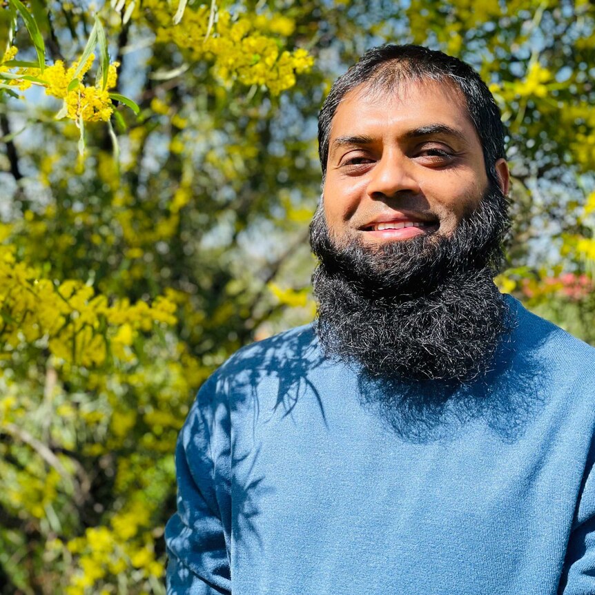 Man in a blue shirt, with a bushy black beard, smiling in front of wattle trees