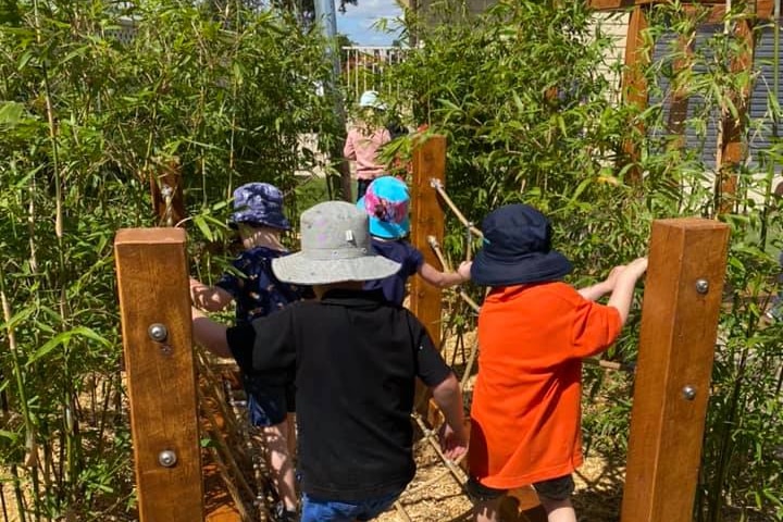 Backs of kids and hats crossing a bridge, with green bamboo trees on either side.