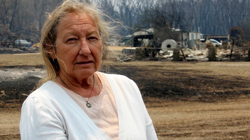 A woman wearing a white cardigan stands in front of charred grass and a burnt building.