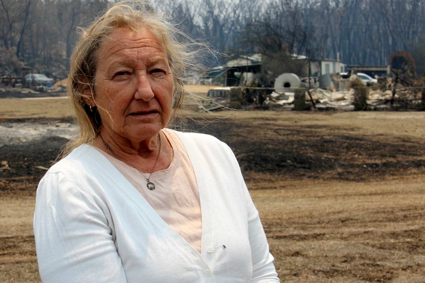 A woman wearing a white cardigan stands in front of charred grass and a burnt building.