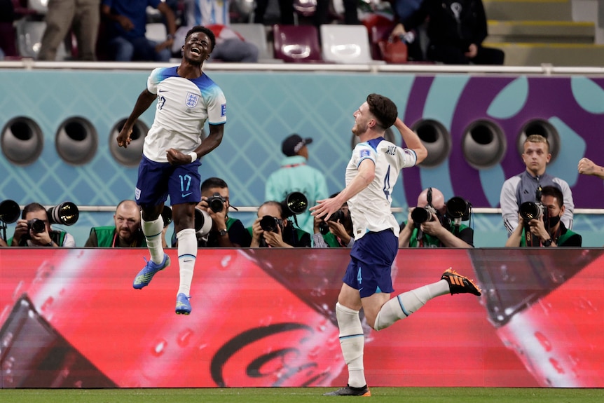 Um jogador de futebol da Inglaterra pula alto em comemoração após marcar um gol na Copa do Mundo enquanto seu companheiro de equipe chuta o ar.