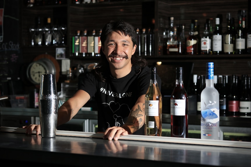 A man with a black moutsache grins behind a bar with spirit bottles and a cocktail shaker on the bar.
