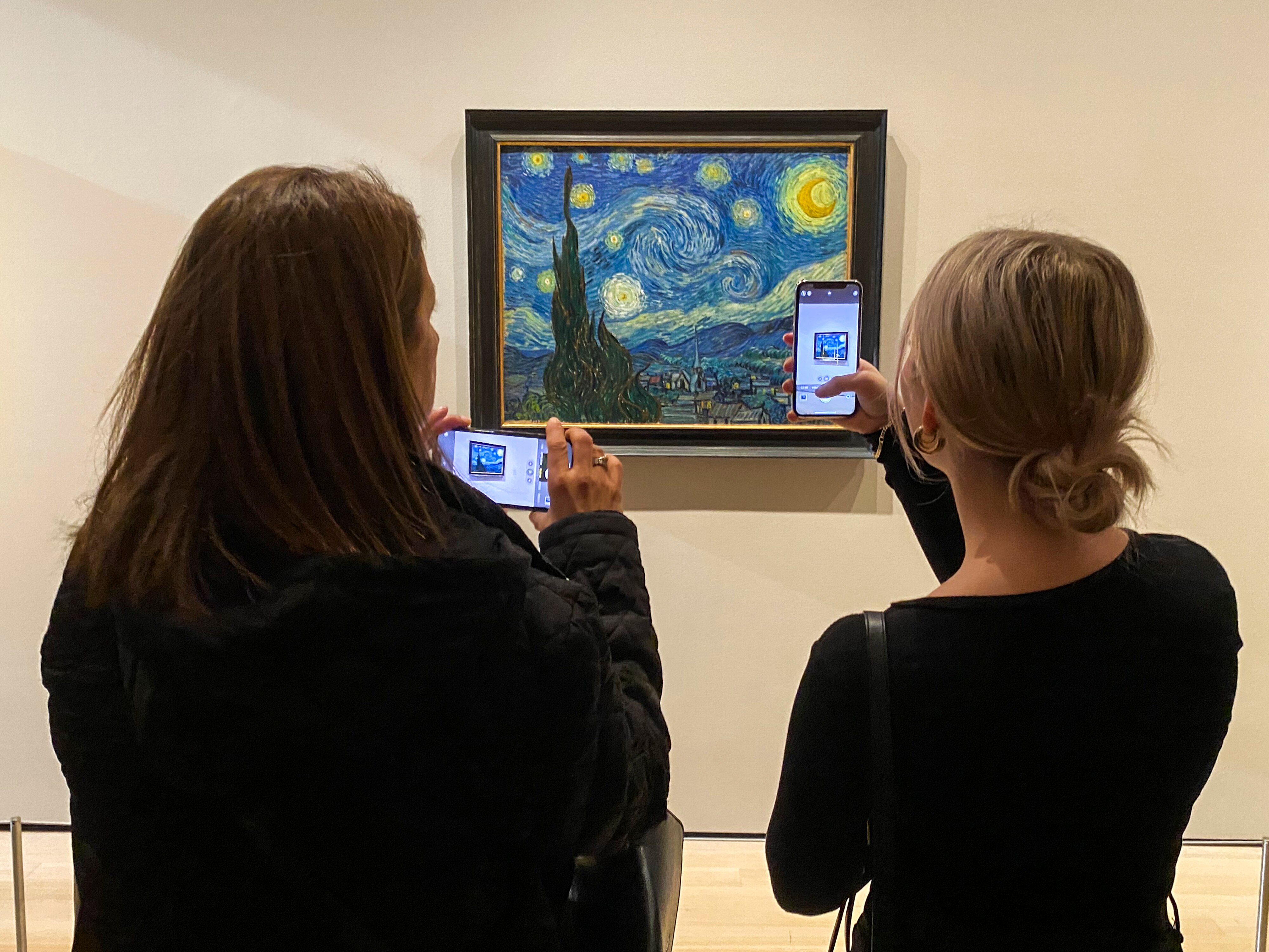 Two people taking a photo of The Starry Night, a painting by Vincent Van Gogh of a town at night.