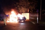 A parked car burns at night as a firefighter approaches on the road, with a brick road in the background showing graffiti.