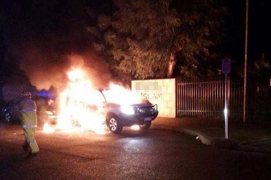 A parked car burns at night as a firefighter approaches on the road, with a brick road in the background showing graffiti.