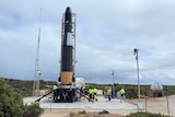Workers in hi-vis jackets working around a rocket standing up ready for take-off, on cement pad, bushes in background.