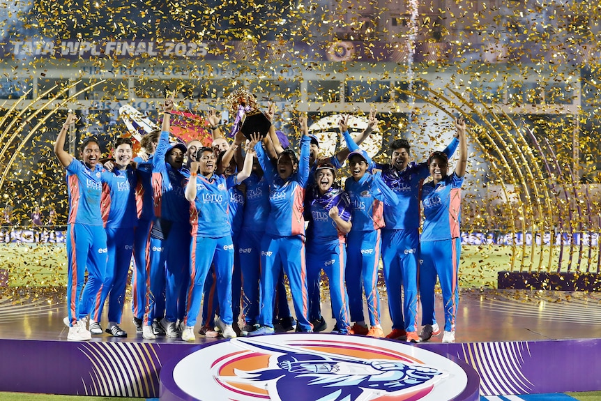 A team of women's cricketers celebrate on a podium with a trophy as confetti falls and a sign in background "WPL FINAL 2023".