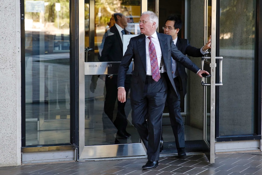 Bret Walker SC walks out a glass door at the High Court, with a phone in his leave hand.