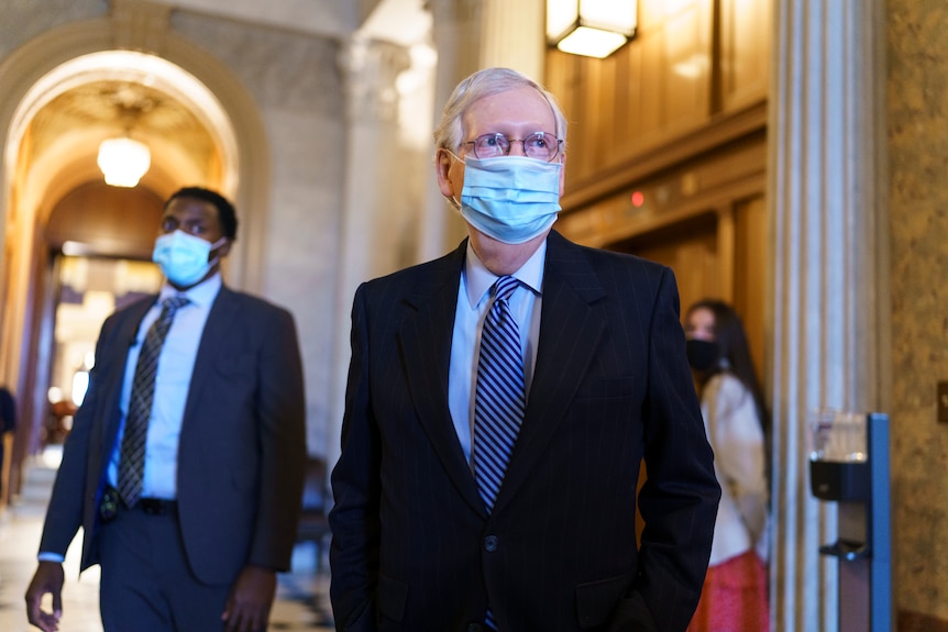 Mitch McConnell walking out of the senate chamber wearing a suit and a blue surgical mask. 