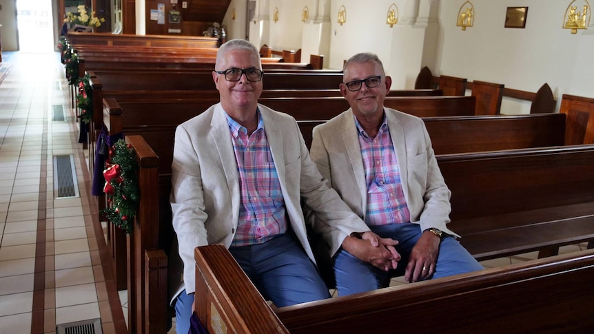 Two men in matching shirts and jackets sit holding hands in a church pew. 