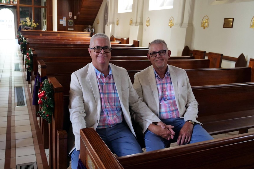 Two men in matching shirts and jackets sit holding hands in a church pew. 