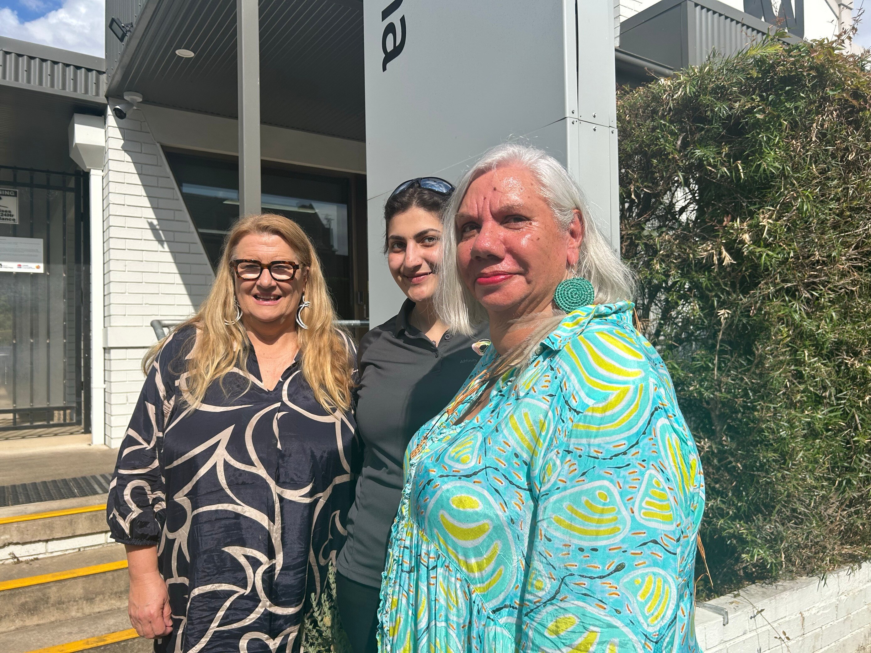 Three women stand on a street, facing the sun