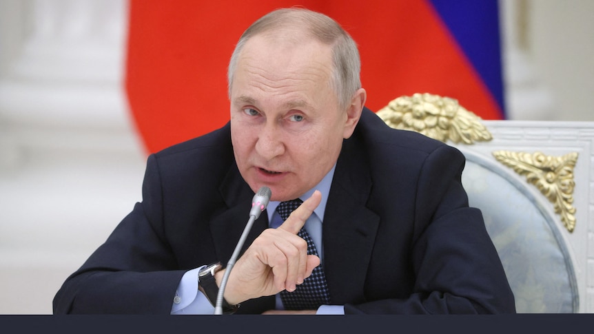 Putin leans forward from where he sits to speak into a microphone on a table, pointing a finger as he does so