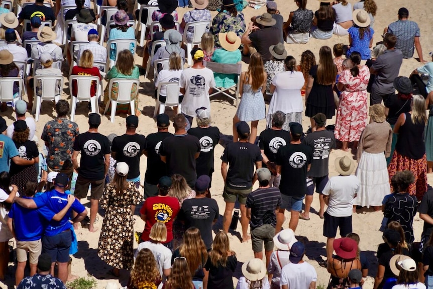people standing on a beach wearing surfing club t-shirts at a funeral. 