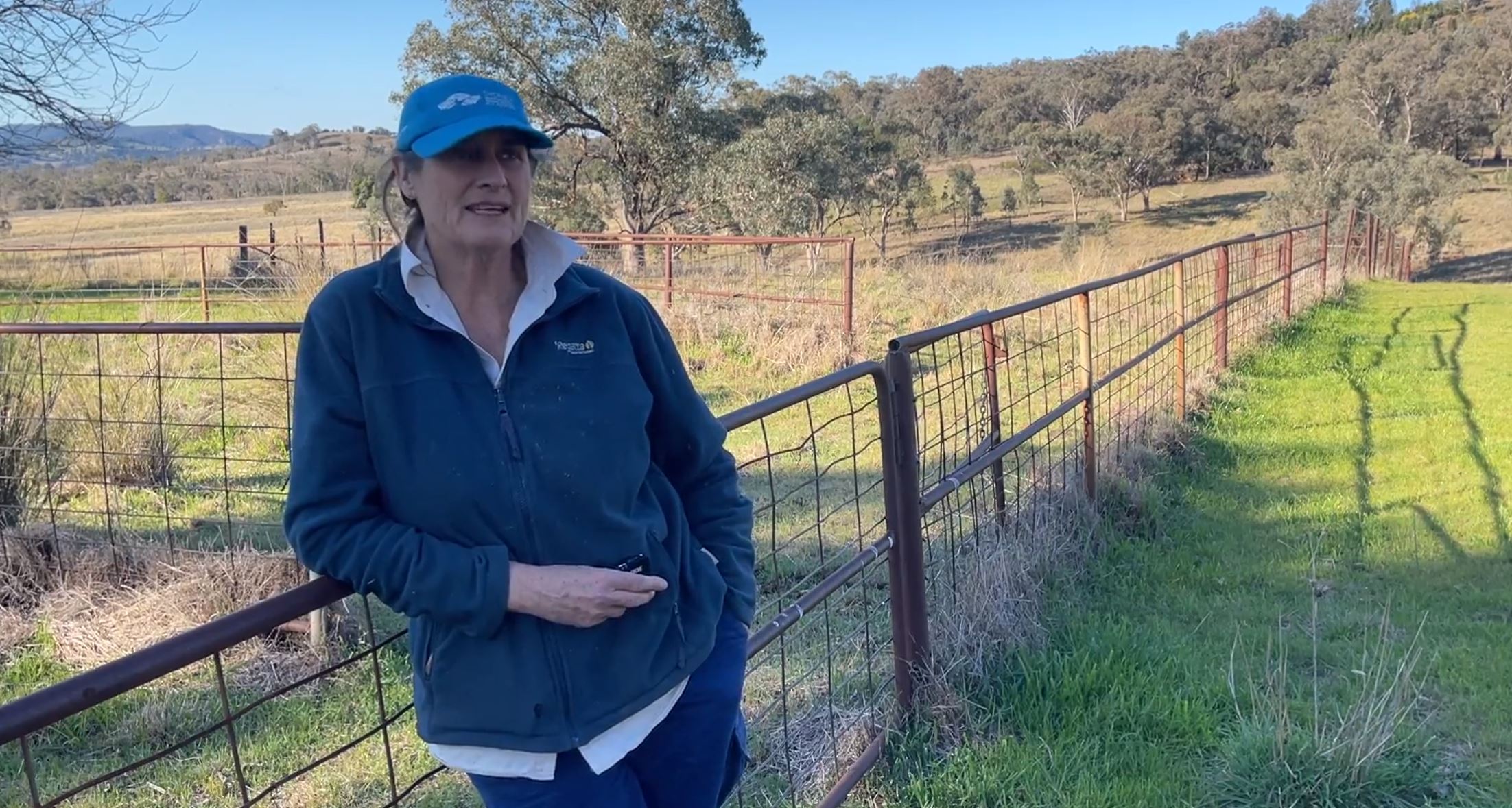 A middle-aged woman in a cap and jumper leans against a fence on a rural property.