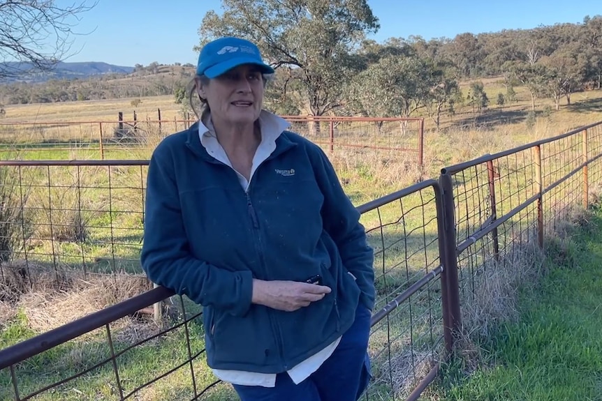 A middle-aged woman in a cap and jumper leans against a fence on a rural property.