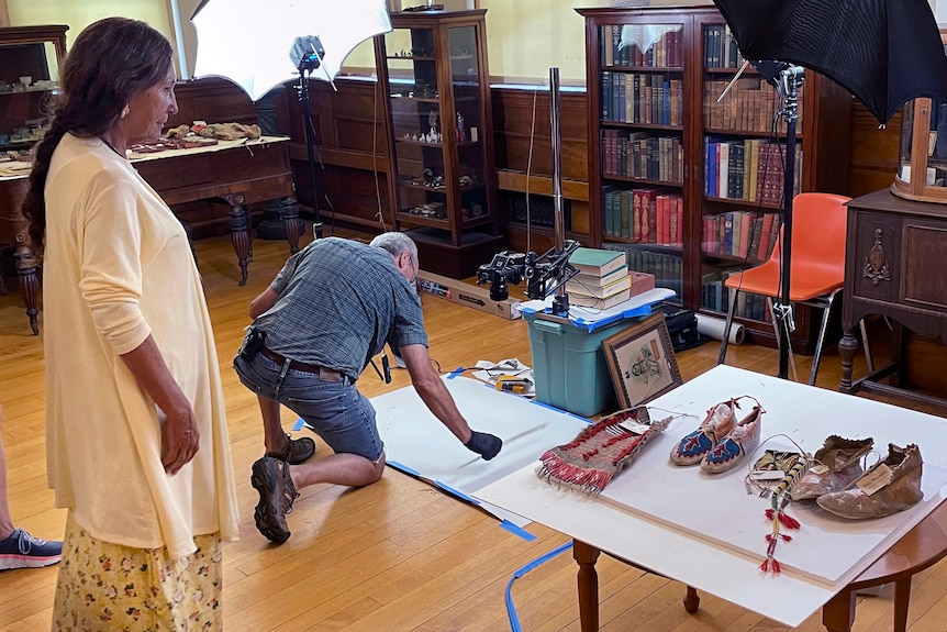 A woman watches a man arrange camera equipment on the floor. Moccassins are set on a table beside her.