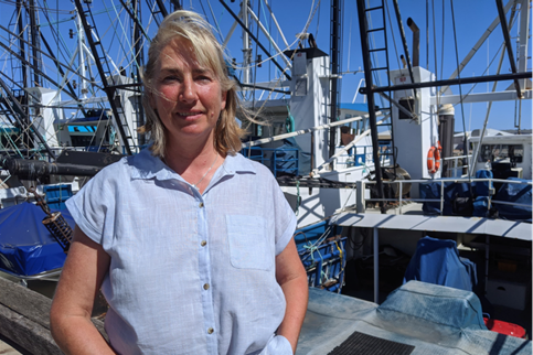 A blonde woman in a blue shirt stand infront of fishing voats.