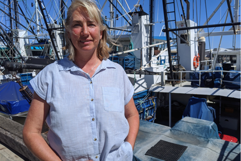 A blonde woman in a blue shirt stand infront of fishing voats.