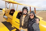 a man and woman with flying helmets stand next to an old yellow plane