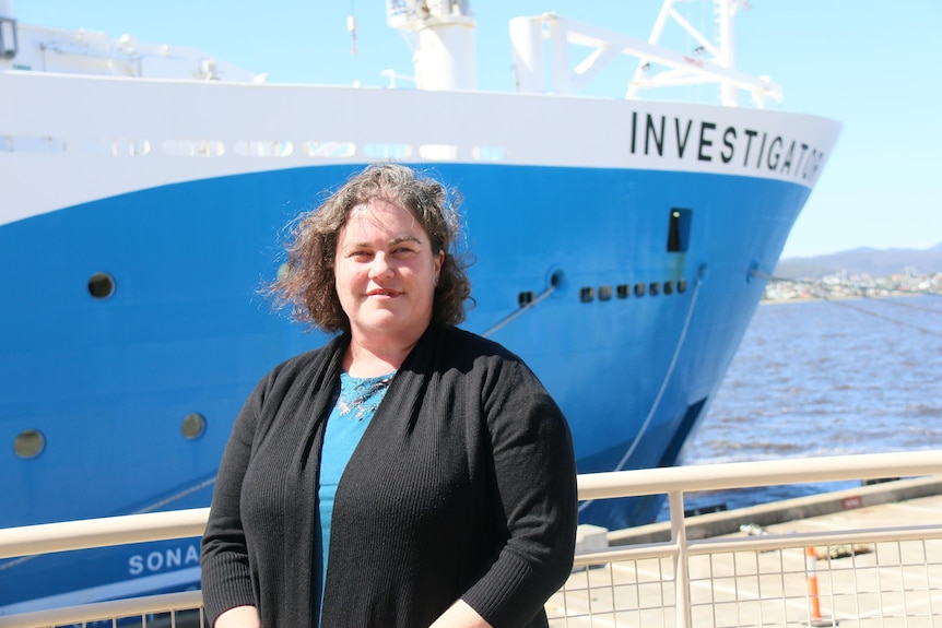 A lady in a blue shirt and black cardigan stands infront of a blue-hulled research ship
