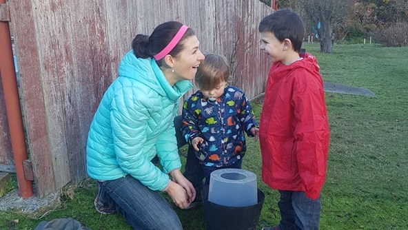 A mother wearing a blue puffer jacket is looking at the mosquito trap with her two-year old and four-year old children.