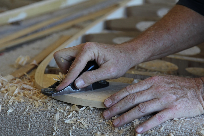 A man's hands on a wooden surfboard frame using a hand plane.