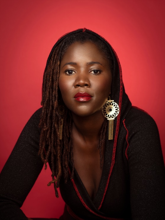 A Black woman with red lipstick and large gold earrings poses for a photo against a red backdrop.
